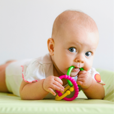 Little baby lying on stomach and looking at camera while teething on plastic rings, representing dentist